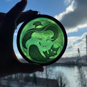Green circular ornament showing a witch with a cat, held up against a blue sky and river cityscape in the background.