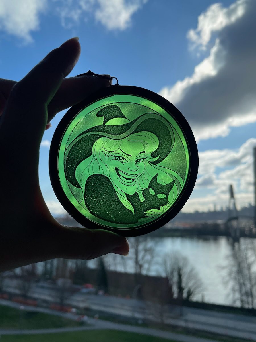 sean-cox-tattoo-glass-art-witch-1-front-sky Green circular ornament showing a witch with a cat, held up against a blue sky and river cityscape in the background.