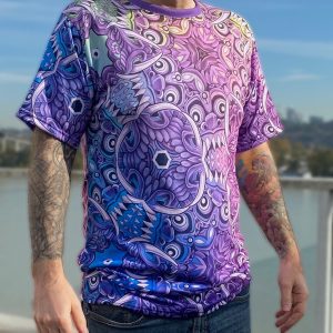 Sean-Cox-Tattoo-Rangdasmagoria-T-Shirt - 1 Man standing outdoors wearing a vibrant, abstract purple-pink-blue T-shirt; tattoos on forearms visible, with a city skyline blurred in the background.